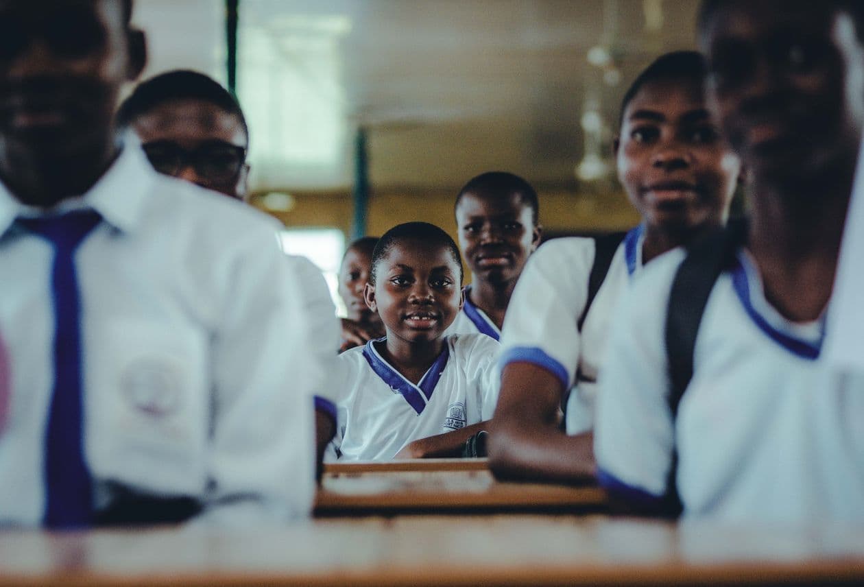 Nigerian secondary school students in white uniforms with blue collars sitting at desks in a classroom, smiling toward the camera