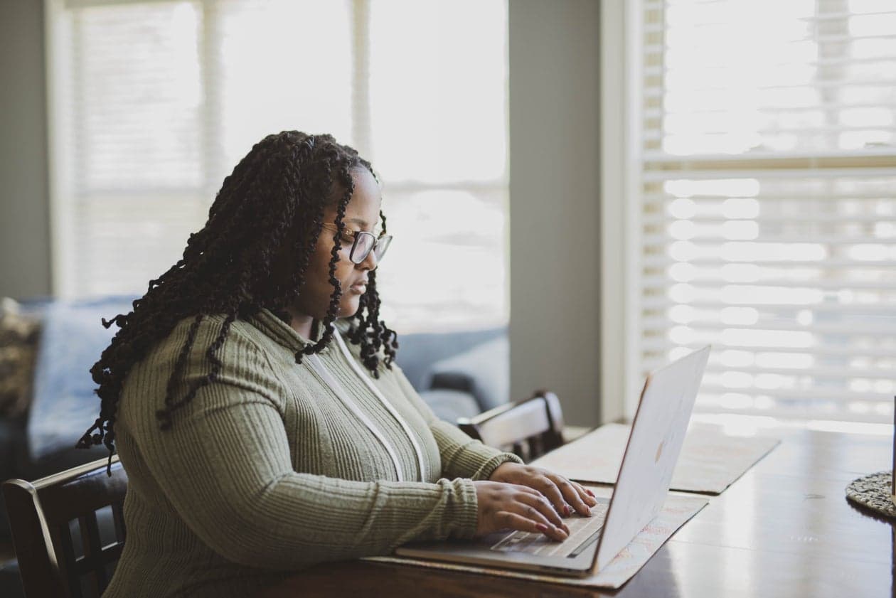 A student with braids and glasses working on a laptop at home